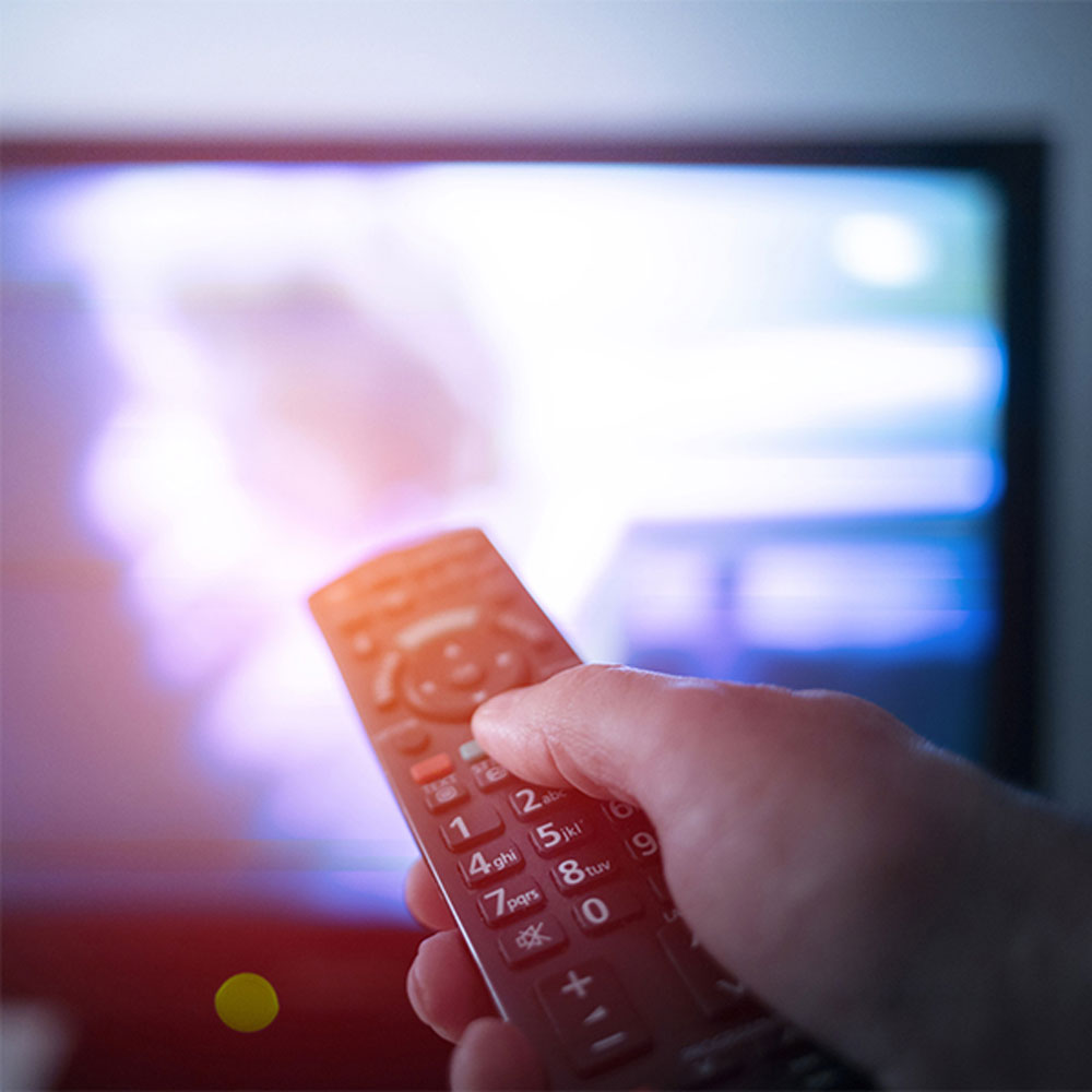 A person points a remote control at a TV with a commercial playing in the background